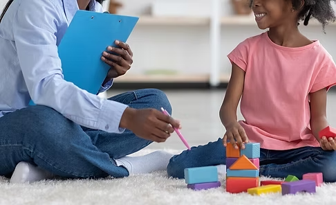 A therapist sitting on the floor with a smiling child, building with colorful blocks during a session
